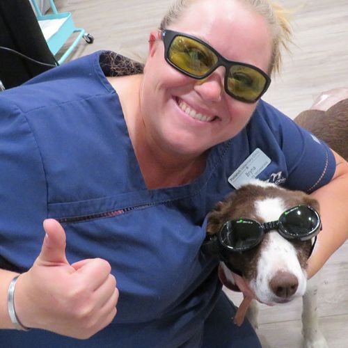 A woman in glasses smiles beside a dog wearing goggles